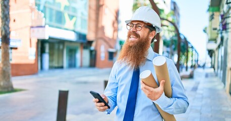 Young redhead architect man with long beard wearing hardhat smiling happy. Holding blueprints using smartphone at street of city.