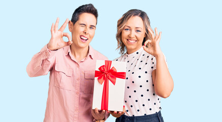 Couple of women holding gift doing ok sign with fingers, smiling friendly gesturing excellent symbol