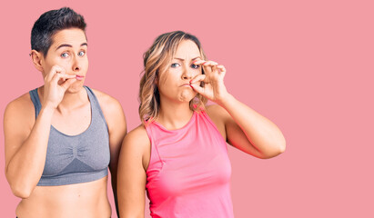 Couple of women wearing sportswear mouth and lips shut as zip with fingers. secret and silent,...
