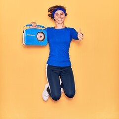 Young beautiful sportswoman listening to music smiling happy. Jumping with smile on face holding vintage radio doing ok sign with thumbs up over isolated yellow background.