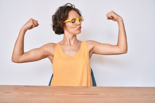 Young hispanic woman wearing casual clothes and glasses sitting on the table showing arms muscles smiling proud. fitness concept.
