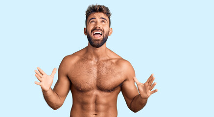 Young hispanic man wearing swimwear shirtless crazy and mad shouting and yelling with aggressive expression and arms raised. frustration concept.