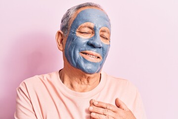 Senior man with grey hair wearing mud mask smiling with hands on chest, eyes closed with grateful gesture on face. health concept.