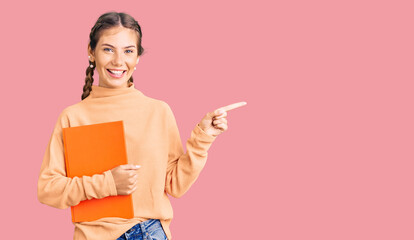 Beautiful caucasian woman with blonde hair holding book smiling happy pointing with hand and finger to the side