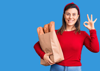 Young beautiful woman holding paper bag with bread doing ok sign with fingers, smiling friendly...