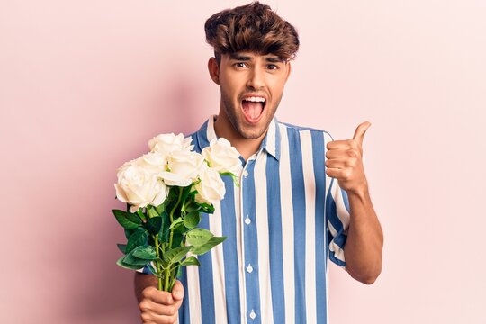 Young hispanic man holding flowers pointing thumb up to the side smiling happy with open mouth