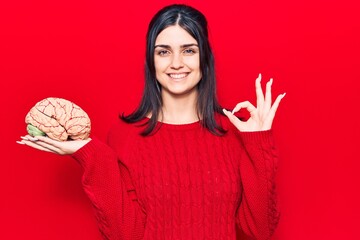 Young beautiful girl holding brain doing ok sign with fingers, smiling friendly gesturing excellent...