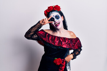 Young woman wearing mexican day of the dead makeup doing peace symbol with fingers over face,...