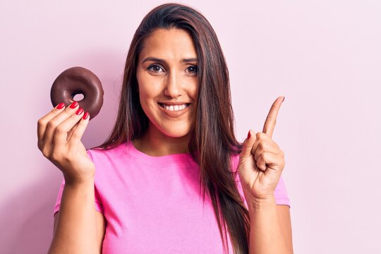 Young beautiful brunette woman holding chocolate donut smiling happy pointing with hand and finger to the side
