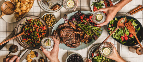 Summer barbeque party. Flat-lay of table with grilled meat, vegetables, salad, roasted potato and peoples hands with glasses of lemon water over white tablecloth, top view, wide composition - 374759243
