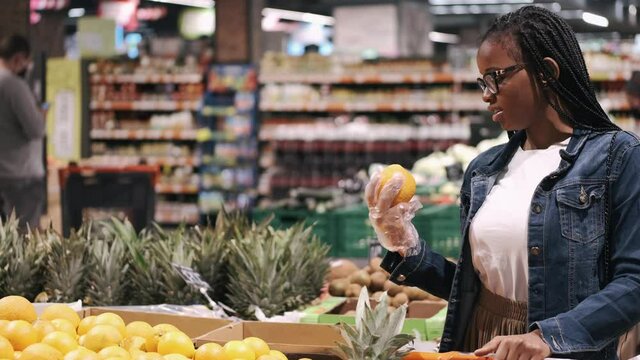 Black young woman is chosing an orange. She is doing shopping in a supermarket. The closeup view.