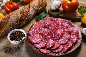 Slices of salami on a clay plate with fresh vegetables and spices on the background of a wooden table. Cold starters.
