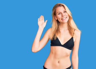 Young beautiful blonde woman wearing bikini waiving saying hello happy and smiling, friendly...