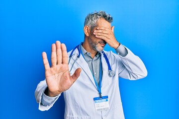 Middle age grey-haired man wearing doctor uniform and stethoscope covering eyes with hands and...