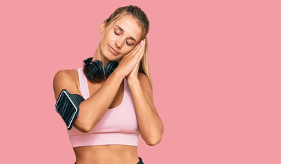 Young blonde woman wearing gym clothes and using headphones sleeping tired dreaming and posing with hands together while smiling with closed eyes.