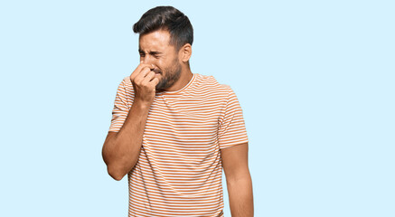 Handsome hispanic man wearing casual clothes smelling something stinky and disgusting, intolerable smell, holding breath with fingers on nose. bad smell