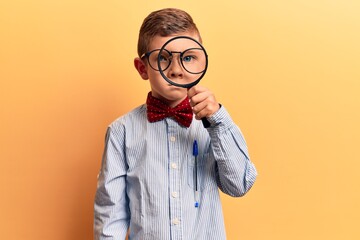 Cute blond kid wearing nerd bow tie and glasses holding magnifying glass thinking attitude and...