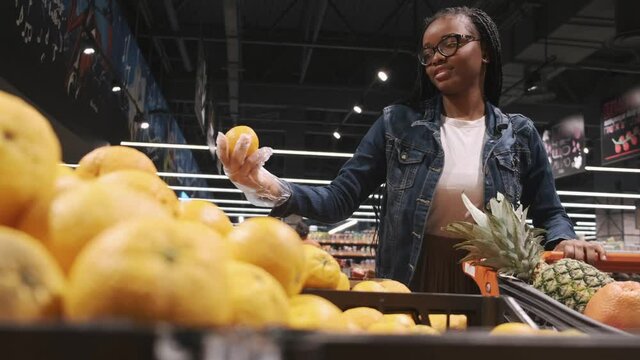 Black young woman is chosing an orange. She is doing shopping in a supermarket. The closeup view.