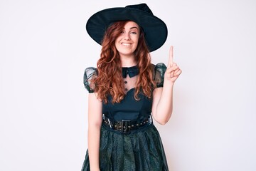Young beautiful woman wearing witch halloween costume showing and pointing up with finger number one while smiling confident and happy.