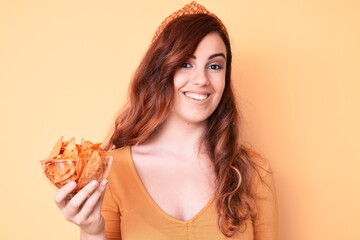 Young beautiful woman holding nachos potato chips looking positive and happy standing and smiling...