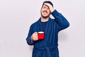 Young hispanic man wearing robe holding coffee stressed and frustrated with hand on head, surprised and angry face