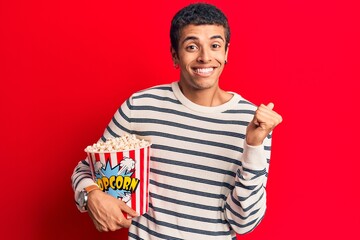 Fototapeta premium Young african amercian man holding popcorn screaming proud, celebrating victory and success very excited with raised arm