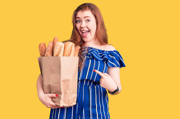 Obraz premium Young beautiful redhead woman holding paper bag with bread smiling happy pointing with hand and finger