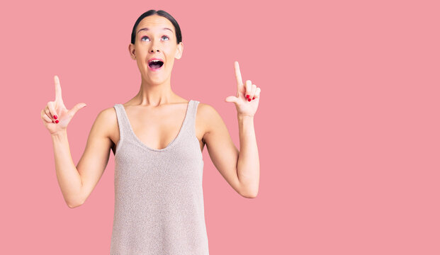 Beautiful brunette young woman wearing casual dress amazed and surprised looking up and pointing with fingers and raised arms.