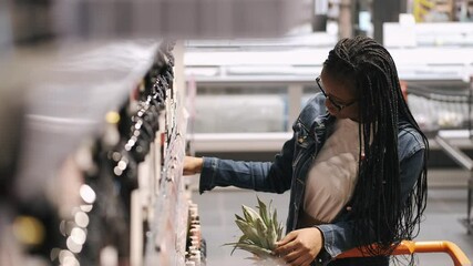 A young black lady is choosing a bottle of wine. The lady is doing shopping in a supermarket. She is at the alcohol department.