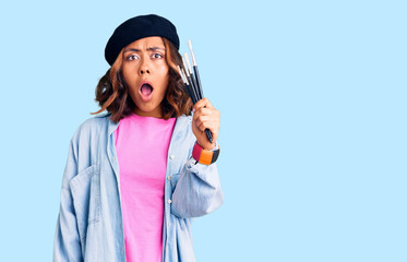 Young beautiful mixed race woman holding paintbrushes scared and amazed with open mouth for surprise, disbelief face