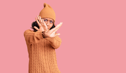 Young beautiful mixed race woman wearing wool sweater and winter hat rejection expression crossing arms and palms doing negative sign, angry face