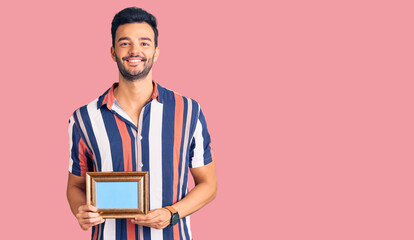 Young handsome hispanic man holding empty frame looking positive and happy standing and smiling with a confident smile showing teeth