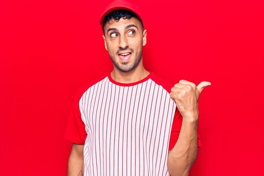 Young hispanic man wearing baseball uniform pointing thumb up to the side smiling happy with open mouth