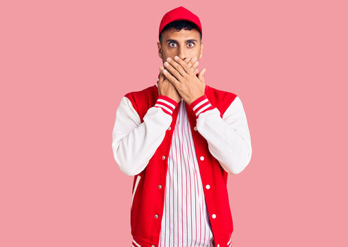 Young hispanic man wearing baseball uniform shocked covering mouth with hands for mistake. secret concept.