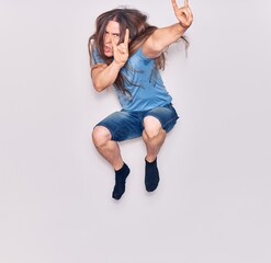 Young handsome man with long hair with aggressive expression wearing casual clothes . Jumping doing heavy metal sign over isolated white background.