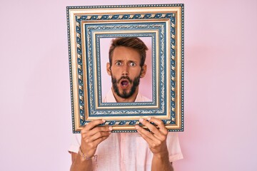Handsome caucasian man with beard holding empty frame in shock face, looking skeptical and...