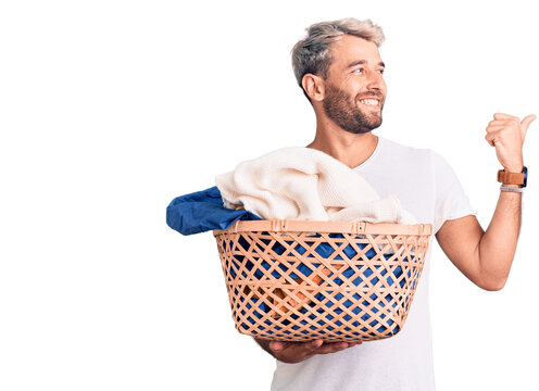 Young handsome blond man holding laundry basket with clothes pointing thumb up to the side smiling happy with open mouth