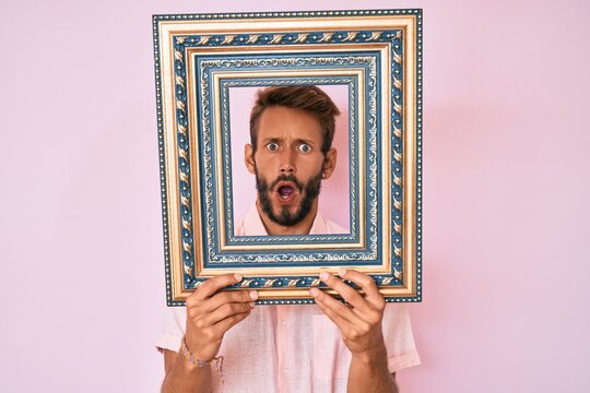 Handsome caucasian man with beard holding empty frame in shock face, looking skeptical and sarcastic, surprised with open mouth