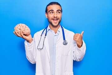 Young handsome man wearing doctor coat holding brain pointing thumb up to the side smiling happy...