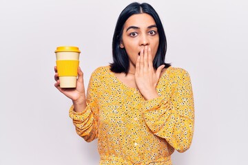Young beautiful latin woman holding takeaway cup of coffee covering mouth with hand, shocked and...