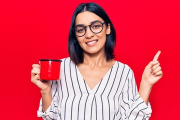 Young beautiful latin woman wearing glasses holding coffee smiling happy pointing with hand and finger to the side