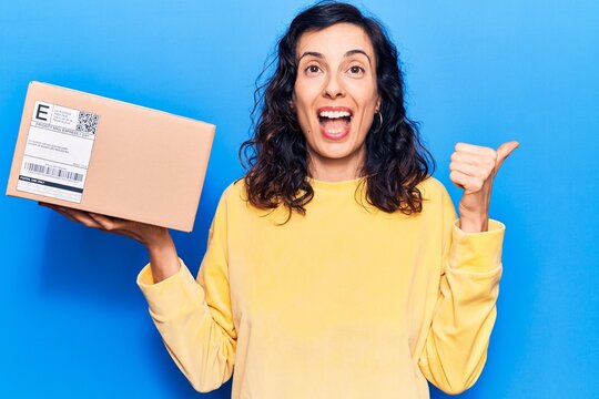 Young beautiful hispanic woman holding delivery package pointing thumb up to the side smiling happy with open mouth