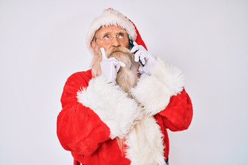 Old senior man wearing santa claus costume speaking on the phone serious face thinking about question with hand on chin, thoughtful about confusing idea