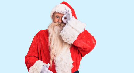Old senior man with grey hair and long beard wearing traditional santa claus costume doing ok gesture with hand smiling, eye looking through fingers with happy face.