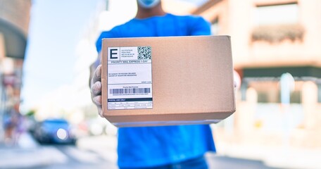 African delivery man wearing courier uniform outdoors wearing coronavirus safety mask holding cardboard parcel