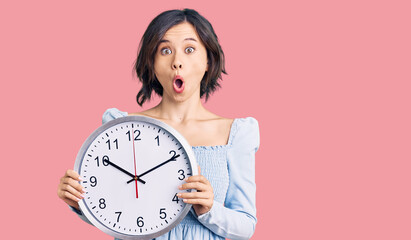 Young beautiful girl holding big clock scared and amazed with open mouth for surprise, disbelief face