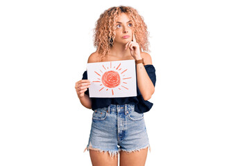 Young blonde woman with curly hair holding sun draw serious face thinking about question with hand on chin, thoughtful about confusing idea