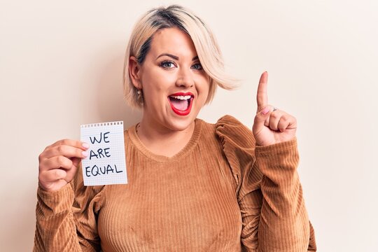 Beautiful blonde plus size woman asking for equality holding paper with we are equal message smiling with an idea or question pointing finger with happy face, number one