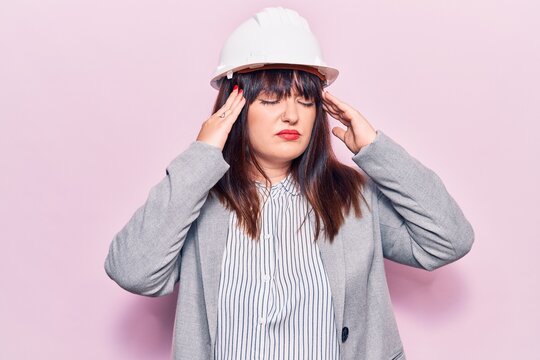 Young plus size woman wearing architect hardhat with hand on head, headache because stress. suffering migraine.