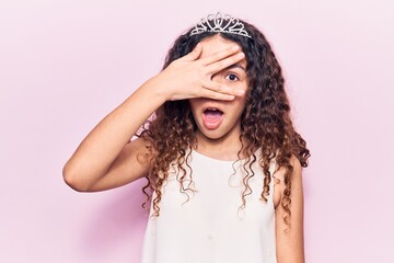 Beautiful kid girl with curly hair wearing princess tiara peeking in shock covering face and eyes with hand, looking through fingers afraid